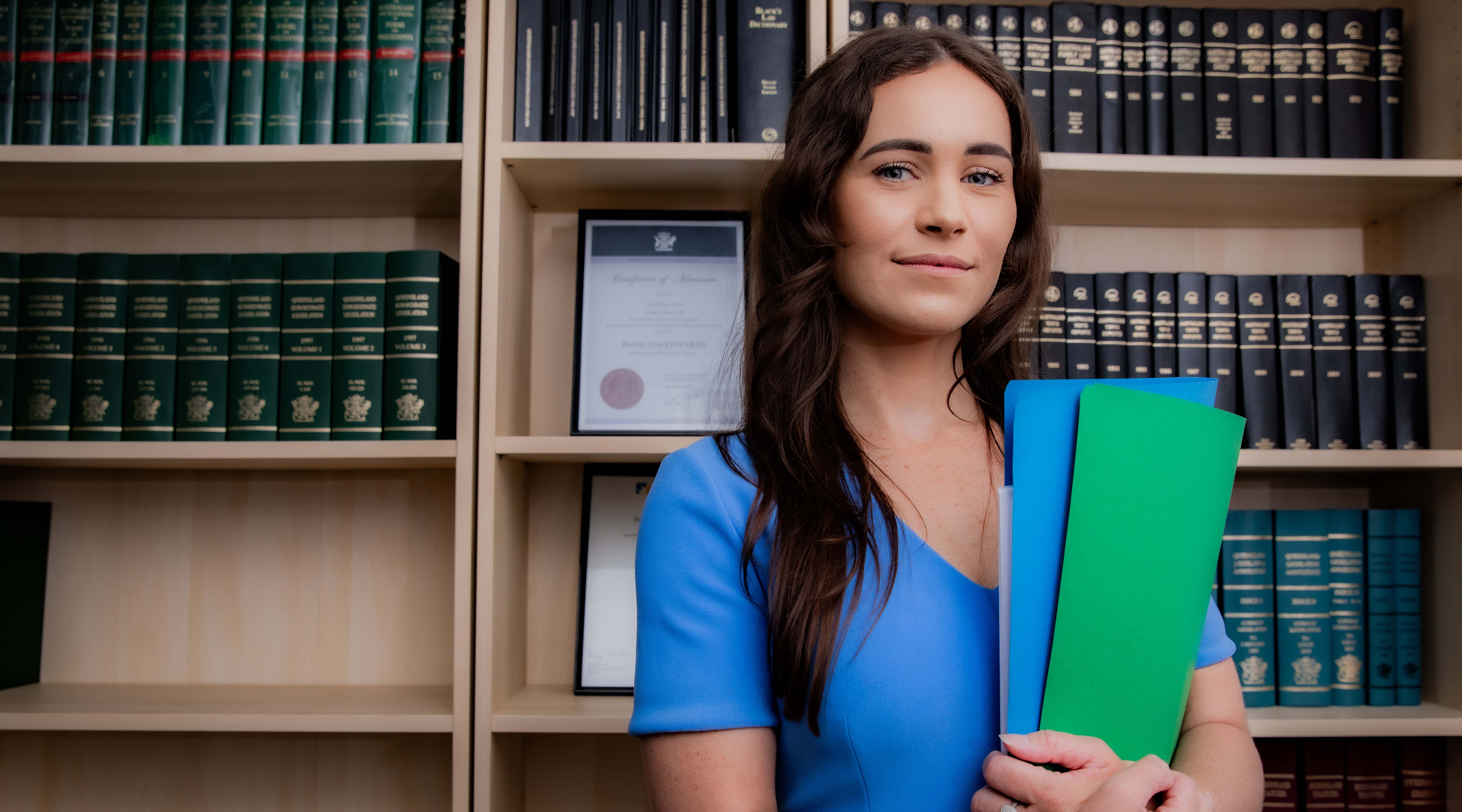 Bridie Edwards, dressed in a blue dress, stands in front of bookshelves filled with law books while holding coloured cardboard folders.