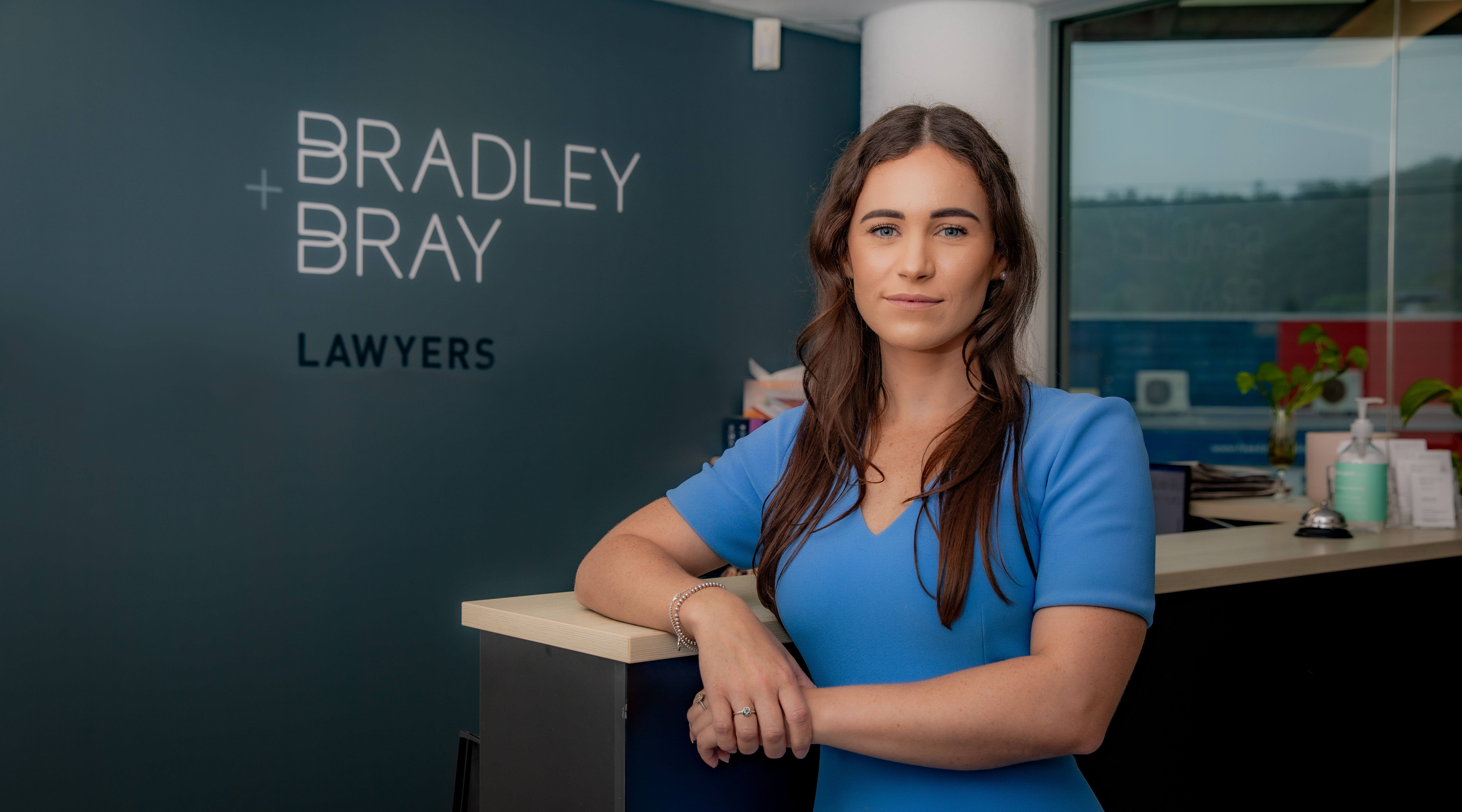 Bridie Edwards, dressed in a blue dress, is leaning on the front desk at Bradley And Bray Lawyers