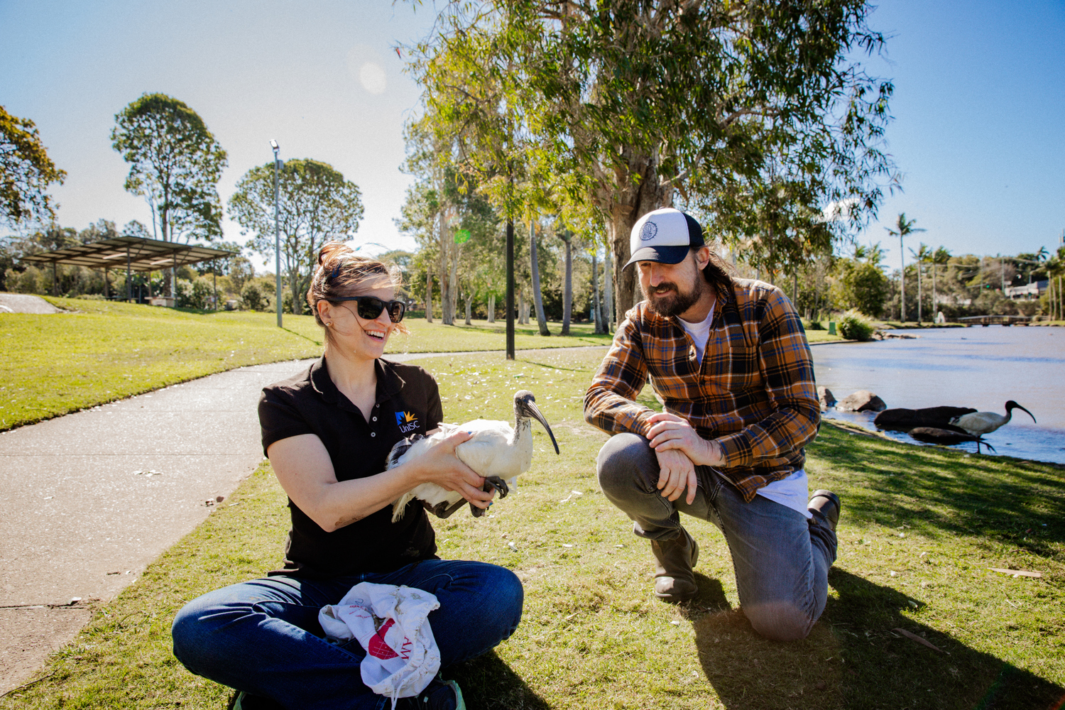 An image of Dr Dominique Potvin sitting cross-legged on the grass, holding a white ibis with both hands, showing it a person in black jeans and a flannelette over-shirt who's crouching next to them. 