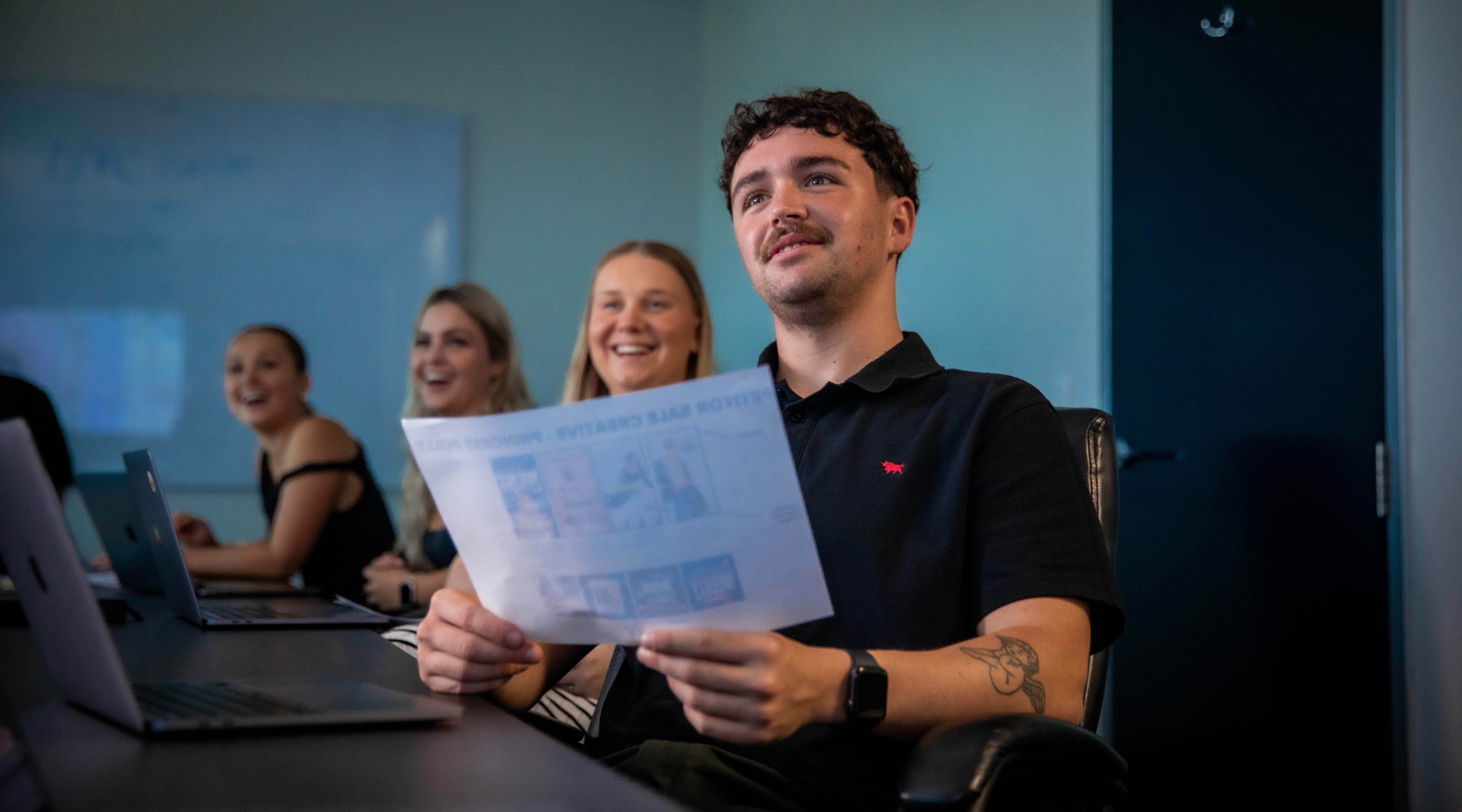 A UniSC graduate is in a business meeting, holding a piece of paper in their hands, watching a speaker who is off camera. 