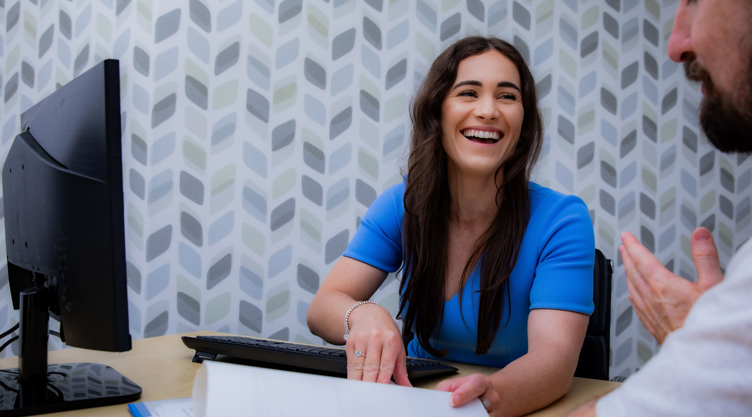 Bridie Edwards, dressed in a blue dress, is sitting at a table in front of a computer and some papers, laughing with someone out of shot.